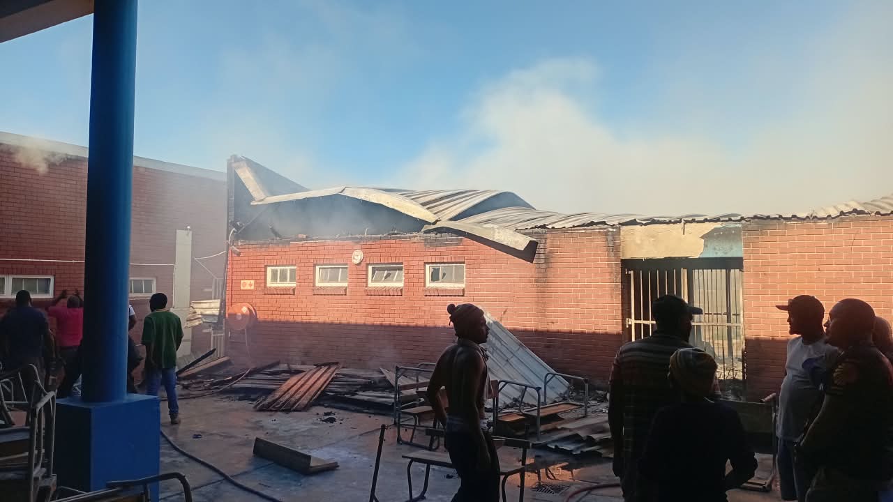 Community members stand in a courtyard looking at a red-brick classroom block whose roof has collapsed, with smoke still rising and burnt desks in the foreground