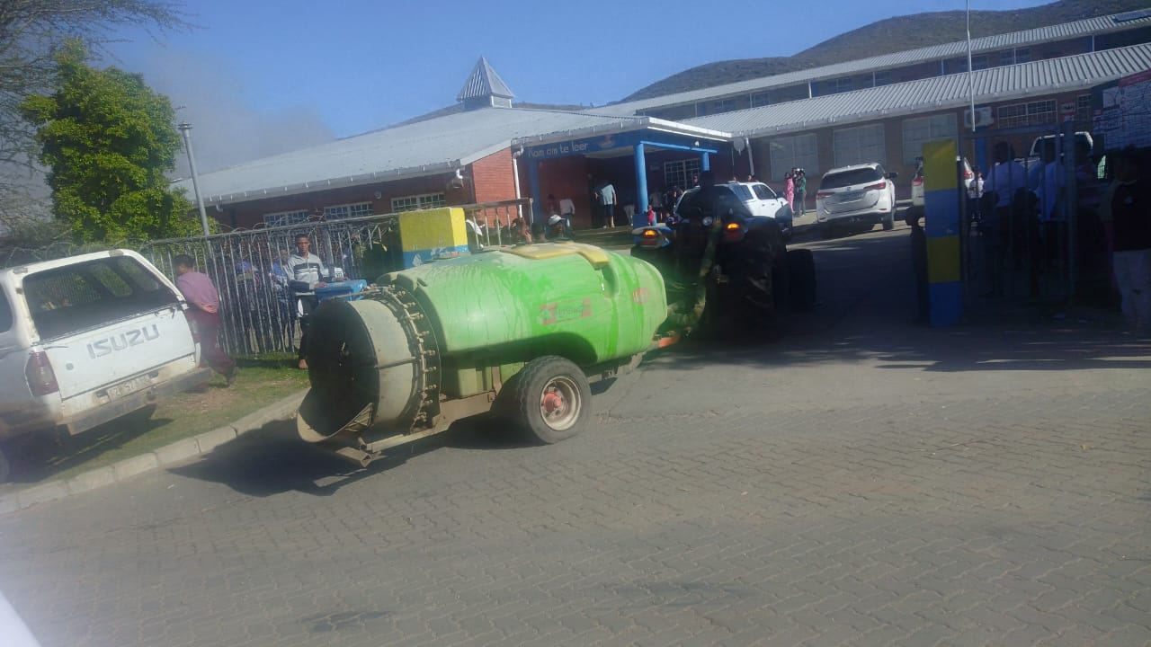 A green orchard sprayer tank towed by a tractor at the gate of Haarlem Secondary School, with smoke visible behind the building
